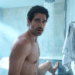 Un hombre con expresión de sorpresa reacciona al agua fría mientras se ducha en un baño moderno iluminado por la luz del sol.