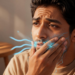 Un hombre de mediana edad con expresión de angustia y malestar se sostiene la mejilla derecha con la mano. Líneas azules onduladas y partículas brillantes emanan de su boca, labios y mejilla, extendiéndose como terminaciones nerviosas por el lado de su cara. Un vaso de agua y una mesa de madera están en el fondo difuminado, en una habitación cálidamente iluminada por el sol.