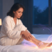 Una mujer sentada en la cama durante la noche se sujeta la pantorrilla con expresión de incomodidad, con un efecto de líneas de luz brillante que rodea su pierna y su pie.