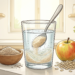 Vaso de agua en el que se mezcla polvo de psyllium con una cuchara, acompañado de un bol con fibra, una manzana y copos de avena sobre una encimera.