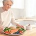 Una mujer mayor sonriente en una cocina luminosa sosteniendo un plato saludable con salmón a la plancha, brócoli, quinua, garbanzos, espinacas y frutos rojos.
