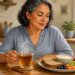 Mujer adulta sentada en una cocina sosteniendo una taza de infusión, junto a un plato con pan, yogur, fruta y una rama de canela sobre la mesa.