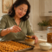 Mujer adulta sirviendo garbanzos tostados en un cuenco, junto a una bandeja llena de garbanzos, un frasco de vidrio, yogur, pera y frutos secos en una cocina.