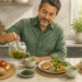 Hombre adulto sirviendo té verde de una tetera en una taza, frente a varias comidas con verduras, pescado, cereales, aguacate, sopa y fruta.