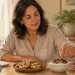 Mujer adulta eligiendo una merienda con yogur, frutos secos, frutos rojos y manzana mientras unas galletas permanecen en un plato sobre la mesa.