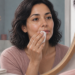 Mujer observa su labio en el espejo mientras aplica una compresa sobre la boca en un baño luminoso, con cuenco con hielo y toallas sobre la encimera.