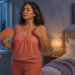 Mujer de mediana edad se abanica junto a una ventana abierta en un dormitorio durante la noche, con gesto de calor y ropa ligera.