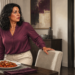Mujer adulta de pie junto a una mesa con un plato de comida, apoyada en una silla y mirando hacia una habitación contigua.