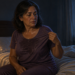 Mujer adulta sentada en la cama por la noche con el pijama húmedo por el sudor, sujetando el cuello de la camiseta junto a una mesilla con lámpara y vaso de agua.