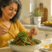 Mujer de mediana edad prepara una ensalada de hojas verdes y verduras en una cocina luminosa, con aceite de oliva, huevo y nueces sobre la mesa.