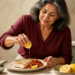 Mujer adulta exprimiendo medio limón sobre un plato con pescado y verduras, con más limones y un pequeño recipiente de sal sobre la mesa.