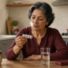 Mujer mayor observa un blíster de comprimidos mientras está sentada en una cocina, con un vaso de agua y un pastillero sobre la mesa.