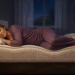 Mujer mayor durmiendo de lado sobre un colchón, con las piernas ligeramente flexionadas y la cabeza apoyada en una almohada, en un dormitorio nocturno.
