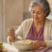 Mujer mayor desayunando un bol con avena, rodajas de plátano y un alimento cremoso blanco, con más plátanos y una taza sobre la mesa.