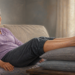 Mujer mayor sentada en un sofá con las piernas elevadas sobre dos almohadas, con varices visibles en las piernas, en una sala iluminada por luz natural.