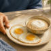 Persona mayor frente a un desayuno con dos huevos, avena con yogur y un vaso de agua, en una mesa iluminada por luz natural.