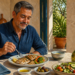 Hombre adulto sentado a la mesa comiendo pescado con arroz y verduras, acompañado de ensalada, aceitunas y una botella de aceite de oliva en un ambiente luminoso.