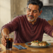 Hombre adulto sentado en una cocina, con una mano sobre un vaso de bebida oscura y la otra sobre un vaso de agua, frente a un plato de comida y un pastelillo.