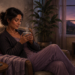 Mujer sentada en un sillón al atardecer, sosteniendo una taza humeante con ambas manos en un ambiente tranquilo, junto a una manta, plantas y una lámpara encendida.