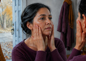 Mujer de mediana edad observando su rostro en un espejo mientras toca suavemente ambas mejillas, en un interior con luz natural y paisaje otoñal al fondo.