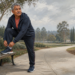 Hombre mayor con ropa deportiva ajustándose una zapatilla con un pie apoyado en un banco antes de hacer ejercicio en un parque.