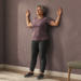 Mujer mayor de cabello gris realizando un ejercicio de postura contra la pared, con los brazos flexionados y las palmas abiertas, en una habitación de casa.