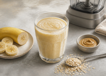 Batido cremoso de plátano en un vaso junto a rodajas de plátano, avena, crema de frutos secos y una licuadora sobre una encimera.