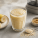 Batido cremoso de plátano en un vaso junto a rodajas de plátano, avena, crema de frutos secos y una licuadora sobre una encimera.