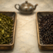 Dos bandejas con hojas de té secas, una de té verde y otra de té negro, colocadas sobre una superficie clara con una tetera al fondo.