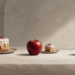 Tres manzanas rojas dispuestas sobre una mesa junto a un pastel grande y una porción pequeña de pastel con cobertura blanca, en una composición comparativa.