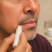 Hombre adulto aplicando un bálsamo en una comisura de los labios, con piel reseca visible alrededor de la boca, frente al espejo de un baño.