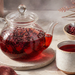 Tetera de vidrio con una infusión roja servida junto a una taza, un vaso de agua y un cuenco con hierbas secas sobre una superficie de cocina.