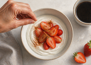 Mano espolvoreando una especia sobre un cuenco de yogur con fresas, acompañado de una taza de café y fresas frescas sobre la mesa.