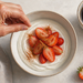Mano espolvoreando una especia sobre un cuenco de yogur con fresas, acompañado de una taza de café y fresas frescas sobre la mesa.