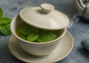 Taza de infusión caliente con hojas frescas de menta, parcialmente cubierta con una tapa de cerámica, junto a más hojas de menta y una tetera.