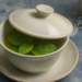 Taza de infusión caliente con hojas frescas de menta, parcialmente cubierta con una tapa de cerámica, junto a más hojas de menta y una tetera.