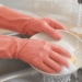Manos protegidas con guantes de goma lavando un plato con una esponja bajo el grifo, junto a un dispensador y una toalla en la cocina.