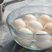 Cuenco de vidrio con varios huevos cocidos enfriándose en agua con cubitos de hielo, junto a un cazo metálico sobre una encimera de cocina.