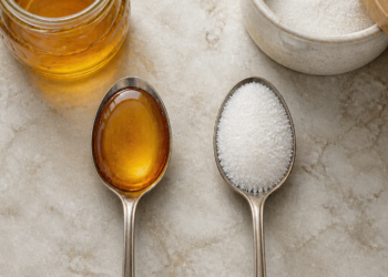 Dos cucharas metálicas sobre una superficie clara, una con miel de color ámbar y otra con azúcar blanco, junto a recipientes con ambos ingredientes.