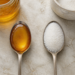 Dos cucharas metálicas sobre una superficie clara, una con miel de color ámbar y otra con azúcar blanco, junto a recipientes con ambos ingredientes.