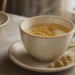 Taza de infusión caliente con pequeñas flores de manzanilla, acompañada de flores secas sobre un plato de cerámica junto a una ventana.