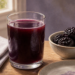 Vaso de bebida de color morado oscuro junto a un cuenco con ciruelas pasas sobre una mesa de madera, una opción asociada al tránsito intestinal y al consumo en ayunas.