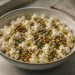 Bol de cerámica con palomitas de maíz, nueces, semillas de calabaza y otros granos, acompañado de un vaso de agua sobre una superficie clara junto a una ventana.