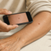 Persona adulta fotografiando con un teléfono móvil una mancha marrón visible en el antebrazo, mientras en la pantalla aparece una ampliación de la lesión cutánea.