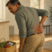 Hombre adulto con gesto de dolor sujetándose la parte baja de la espalda en una cocina luminosa, con una zona rojiza marcada sobre el área lumbar.