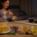 Varias tazas y vasos con infusiones sobre una mesa, con flores y hierbas visibles, mientras una mujer mayor prepara una bebida caliente por la noche en una habitación con la cama al fondo.