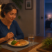 Mujer mayor cenando pescado, verduras y cereales integrales en una mesa durante la noche, con un dormitorio iluminado al fondo.