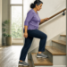 Mujer mayor subiendo una escalera en casa mientras se sujeta del pasamanos, con ropa cómoda y zapatillas deportivas.