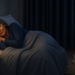 Mujer mayor durmiendo de lado en la cama durante la noche, arropada con una manta azul en un dormitorio tenue con mesita de noche, lámpara y un teléfono móvil al lado.