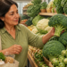 Mujer adulta eligiendo un brócoli en la sección de verduras de un mercado, rodeada de coliflor, coles de Bruselas y otras hortalizas verdes, con una bolsa reutilizable con alimentos.